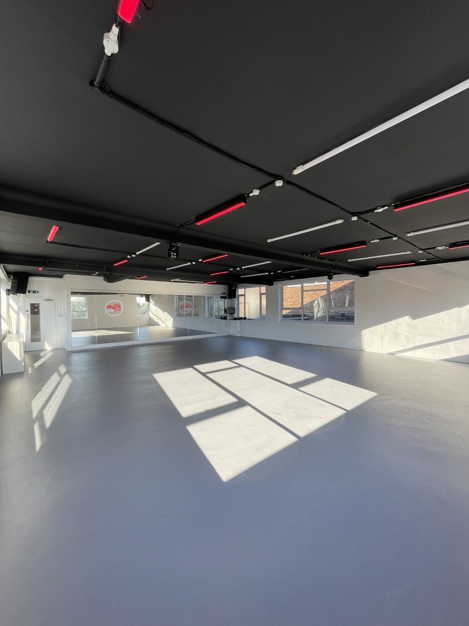 The main studio: a wide grey dance floor in afternoon sun, a full-length mirror wall at the far end, and a black ceiling lined with red and white strip lights.